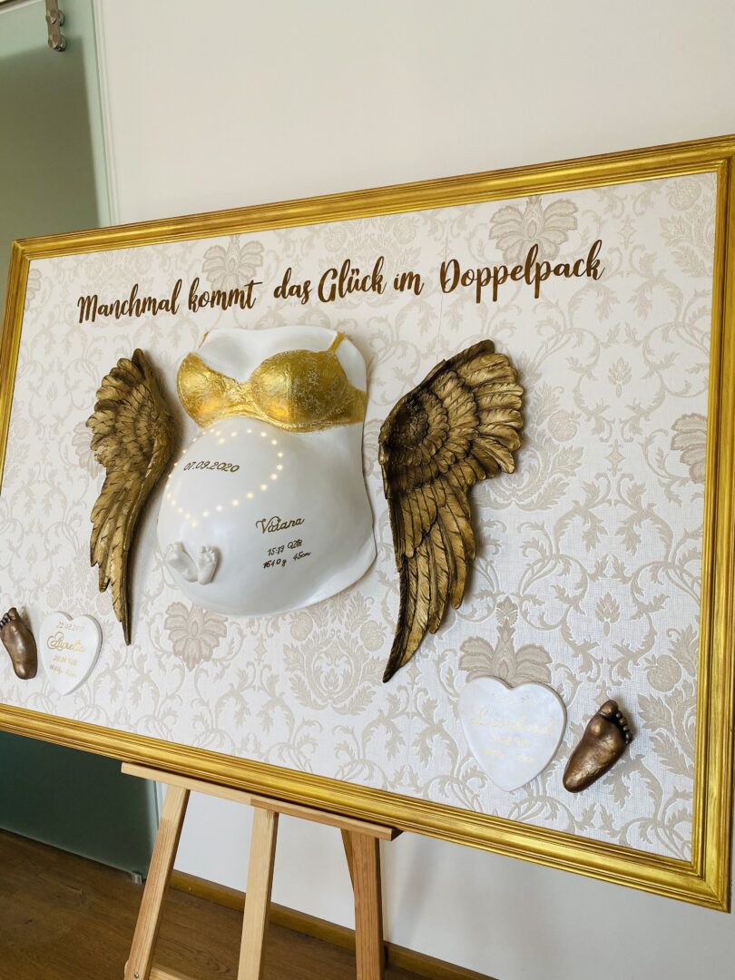 Großer Babybauch-Gipsabdruck im Rahmen mit goldenen Flügeln und integrierten Babyfüßchen