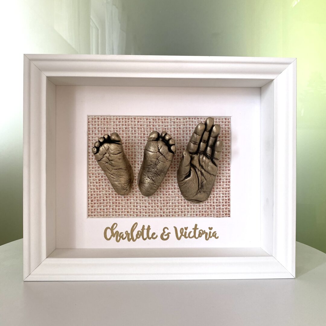 Babyfüße und die Hand der großen Schwester als detailgetreue Gipsabformungen in Antikgold, präsentiert in einem weißen Bilderrahmen