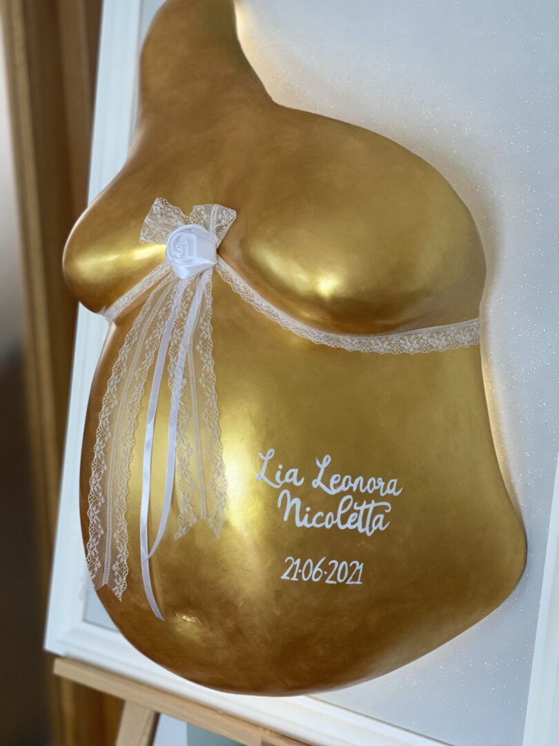 Goldener Babybauch Gipsabdruck mit weißer Spitzenverzierung, Schleife und persönlicher Beschriftung