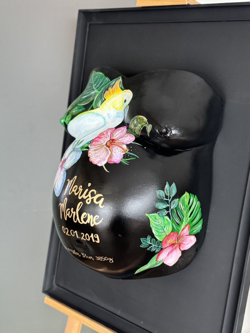 Babybauch Gipsabdruck mit Blumen und Papagei in schwarzem Rahmen